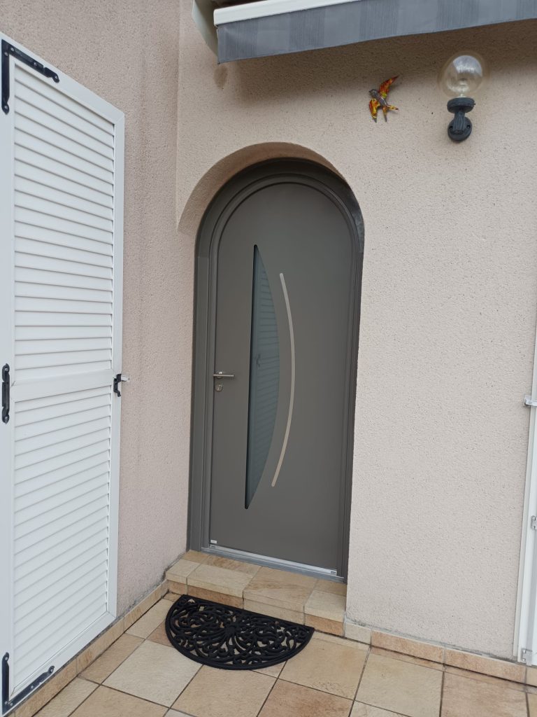 Porte d’entrée cintrée grise avec vitrage étroit courbe et décor argenté, intégrée à une façade beige.