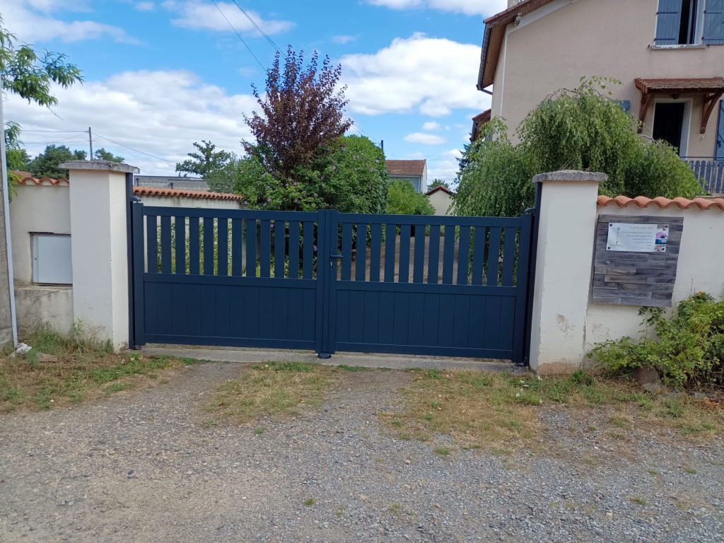 Portail battant bleu foncé à deux vantaux, avec soubassement plein et barreaux verticaux, entre deux piliers blancs.