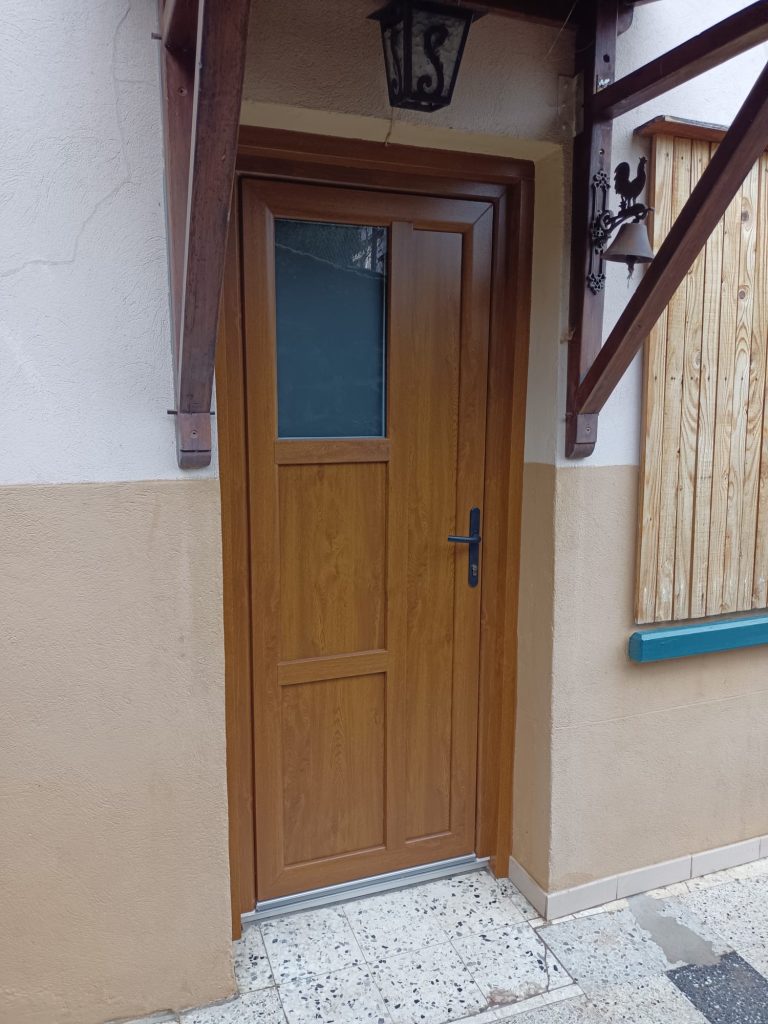Porte d’entrée aspect bois avec vitrage rectangulaire en partie haute et poignée noire, sur une façade beige.