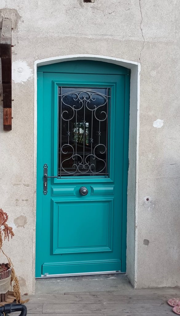 Porte d’entrée turquoise avec vitrage à grille décorative noire, panneaux moulurés et bouton central.