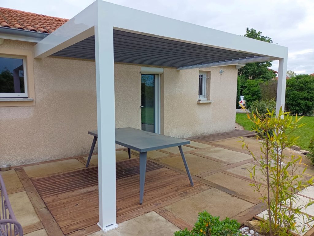 Pergola à structure blanche et toiture à lames grises, adossée à une maison au-dessus d’une terrasse avec une table.