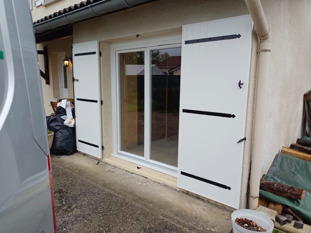 Volets battants blancs à pentures noires, ouverts de chaque côté d’une baie vitrée sur une façade beige.