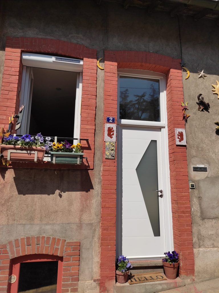 Porte d’entrée blanche avec vitrage géométrique allongé et imposte vitrée, dans un encadrement en briques rouges.