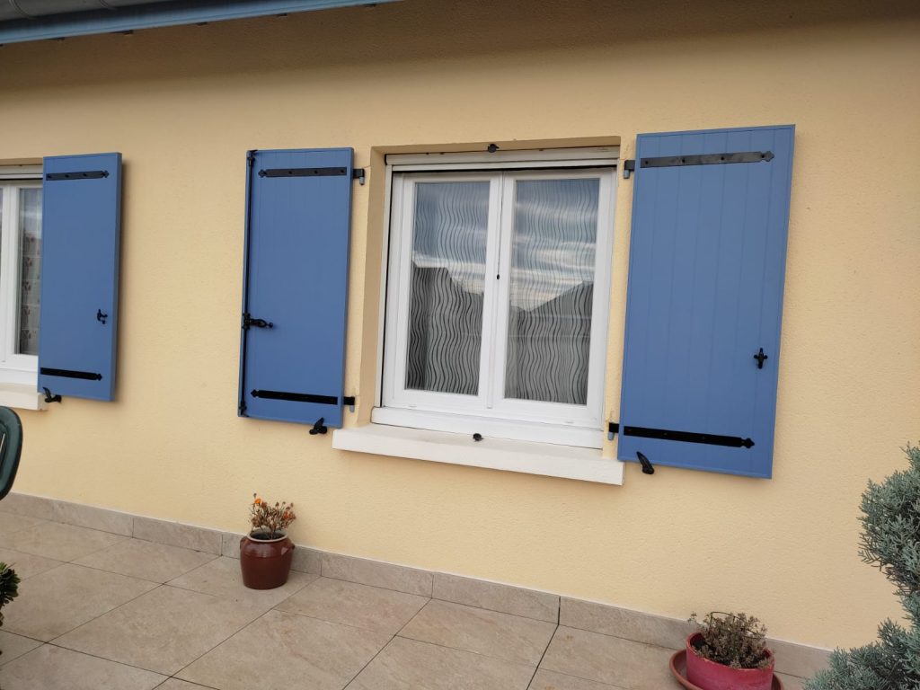 Volets battants bleus à pentures noires, ouverts autour de fenêtres blanches sur une façade jaune clair.