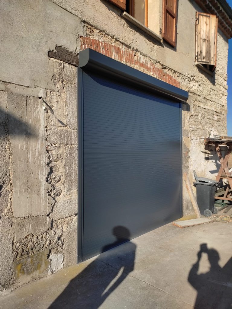 Porte de garage enroulable gris anthracite avec coffre extérieur, installée sur une façade ancienne en pierre et en briques.