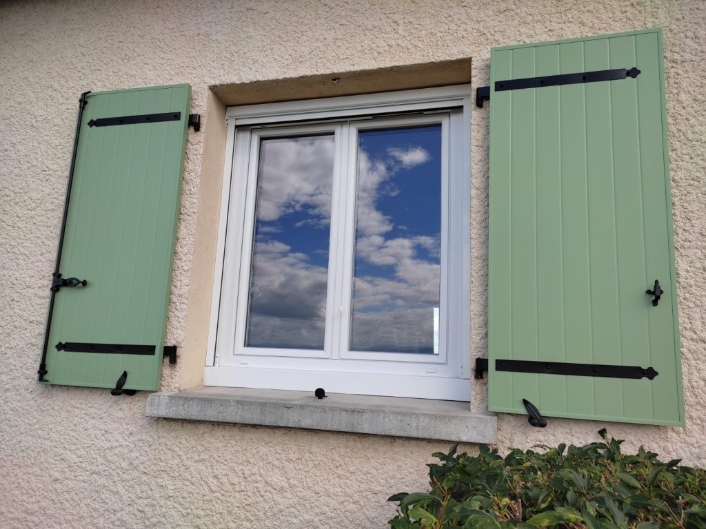 Volets battants vert clair à pentures noires, ouverts de chaque côté d’une fenêtre blanche sur une façade beige.