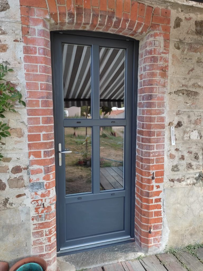 Porte d’entrée gris anthracite à quatre vitrages et soubassement plein, dans un encadrement cintré en briques rouges.